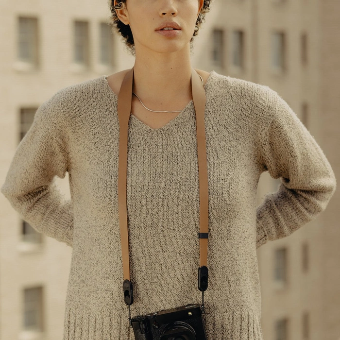Person wearing a beige sweater with a Tan Form Leather Strap over their shoulder, standing in front of a blurred building.