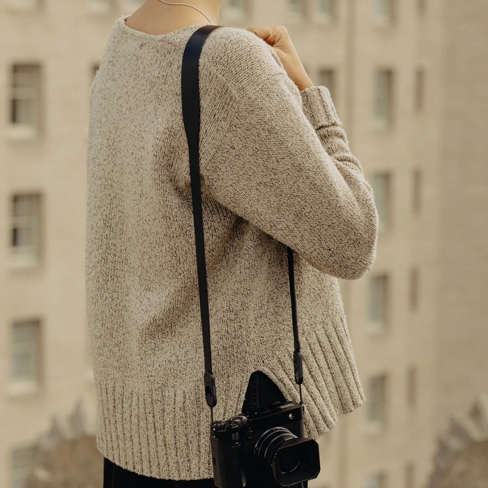 Person wearing a beige sweater with Black Form Leather Strap Standard over the shoulder with a blurred building background