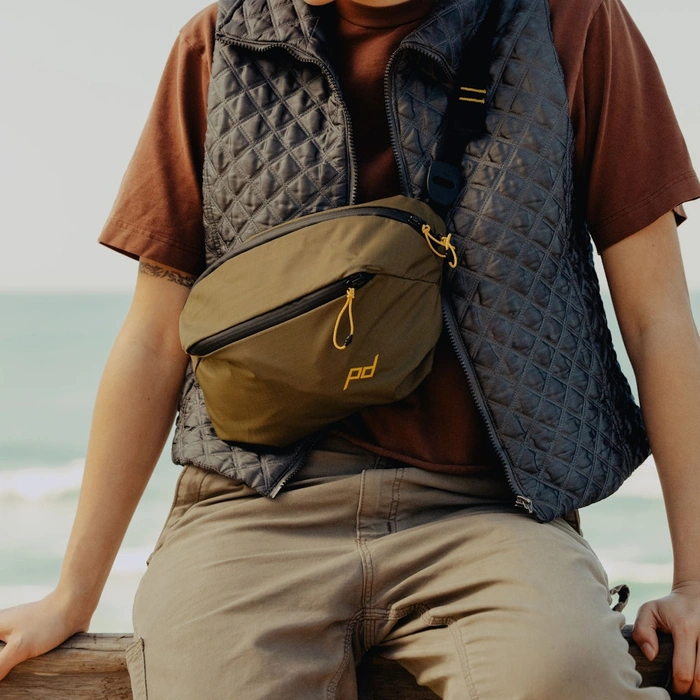 Sitting by the ocean wearing the Outdoor Sling 7L Kelp